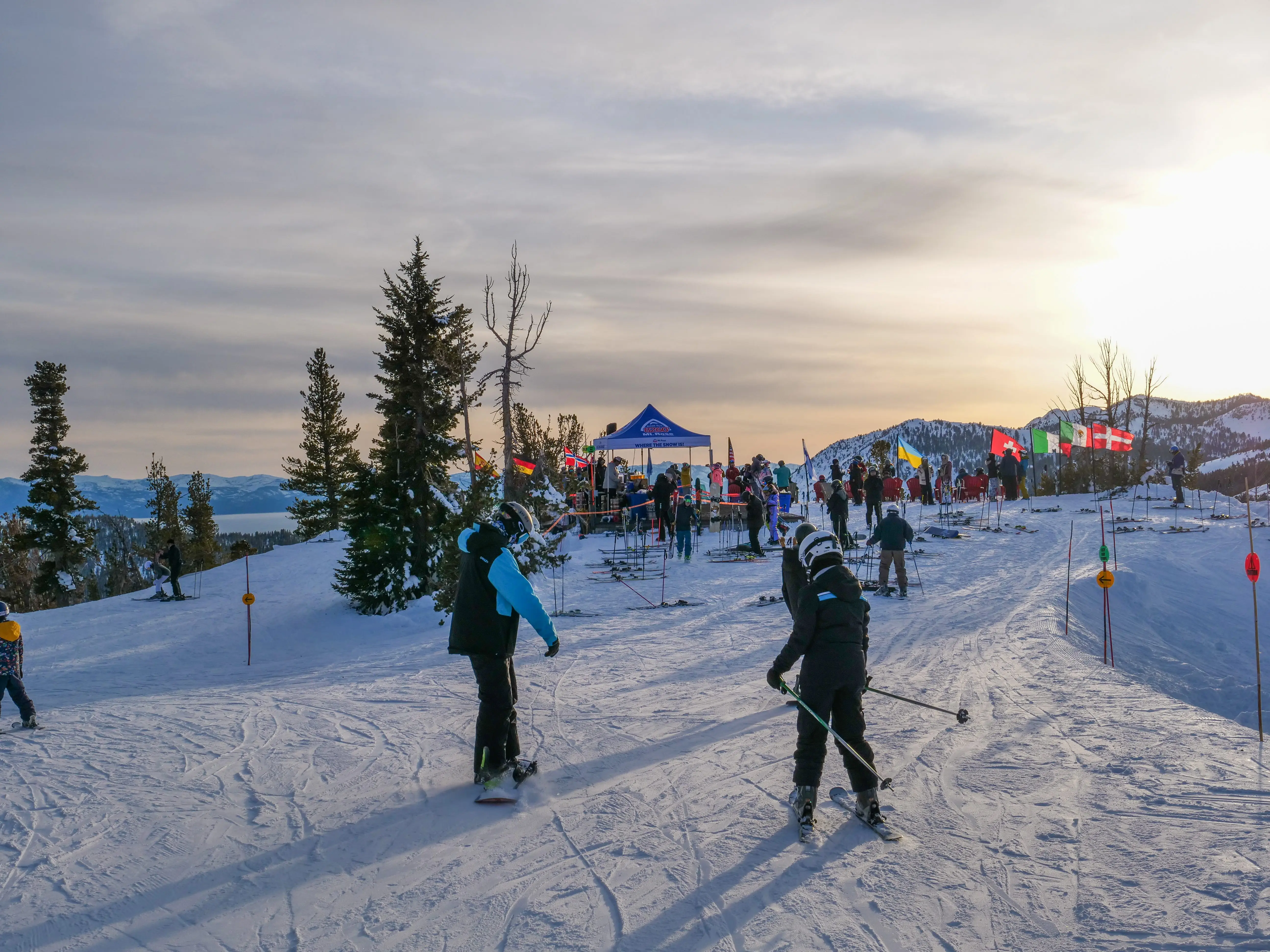 Skiers in winter gear gather at the top of a snowy slope near a tent with colorful flags, surrounded by trees and distant mountains under a cloudy sky at sunset. Mt. Rose Ski Tahoe