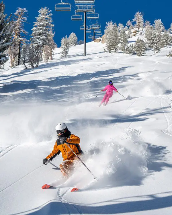 Two skiers in bright orange and pink outfits glide down a snowy mountain slope at Ski Tahoe, kicking up powder as snow-covered trees and empty ski lifts rest under a clear blue sky. Mt. Rose Ski Tahoe