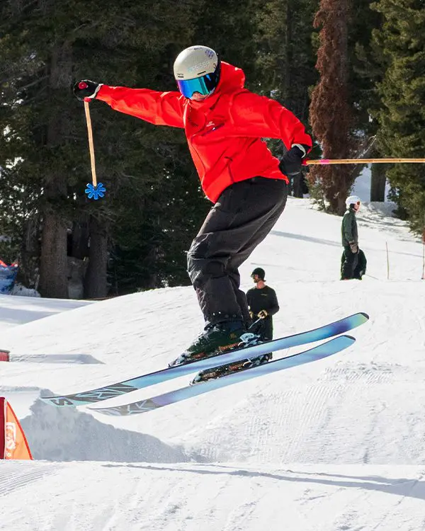 A skier in a red jacket and helmet jumps off a snowy Ski Tahoe slope, holding a ski pole in one hand, with trees and two people standing in the background. Mt. Rose Ski Tahoe