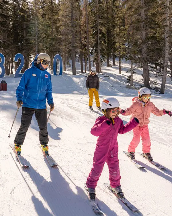 Two adults and two children wearing helmets and ski gear ski down a snowy Ski Tahoe slope among tall trees. The children, in bright pink suits, lead the way. Large blue numbers, 8200, are visible in the background. Mt. Rose Ski Tahoe