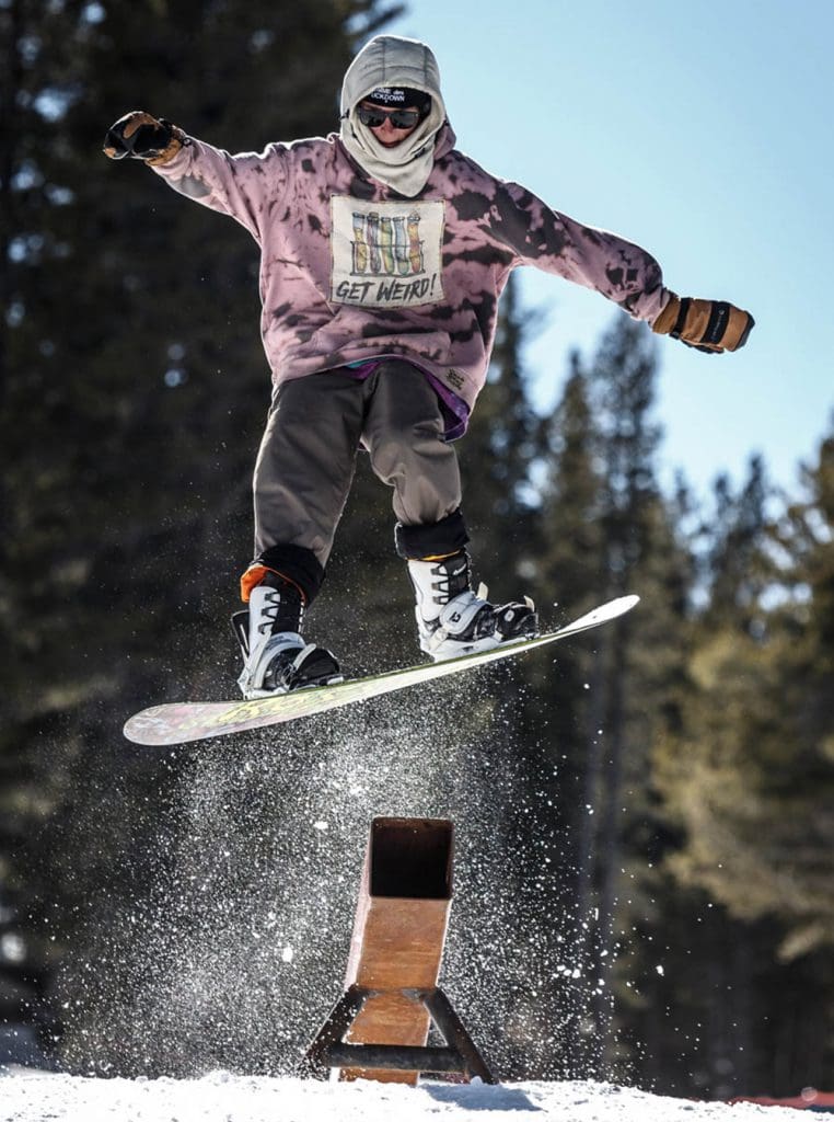 A snowboarder wearing a hoodie, gloves, and goggles performs a jump trick over a metal rail in a snowy outdoor area with trees in the background. Snow sprays around the snowboard as they soar through the air, showing skill and balance. Mt. Rose Ski Tahoe