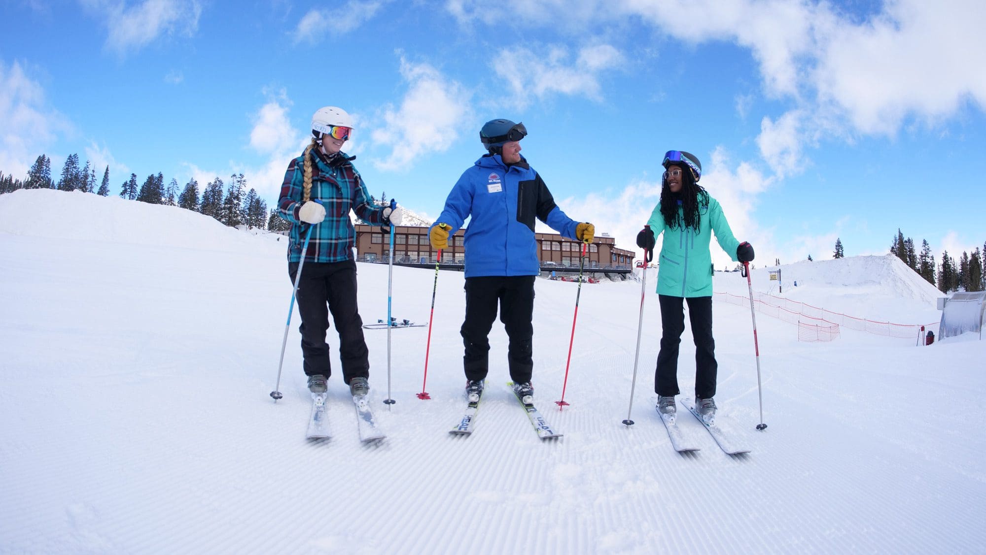 A group of four people, including children and adults, are skiing down a snowy slope on a sunny day. They are all wearing winter gear, including helmets, jackets, and ski boots. There are colorful markers on the snow from the best kids ski lessons, tall pine trees, and a clear blue sky in the background. Mt. Rose Ski Tahoe