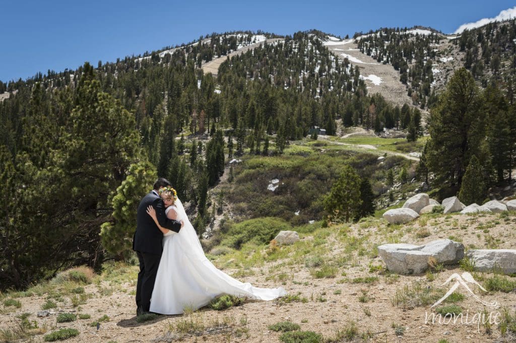 A bride and groom embrace in a mountainous landscape with scattered trees and patches of snow. The bride wears a white dress with a long train and floral headpiece while the groom wears a black suit. The clear blue sky forms the perfect backdrop for their wedding day. "imonie" is written in the bottom right. Mt. Rose Ski Tahoe