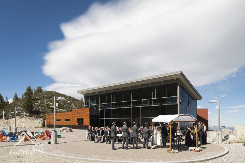 A beautiful wedding ceremony is taking place outdoors in front of a modern glass building surrounded by scenic mountains. Guests are seated facing the couple and officiant under a white canopy. The sky is clear with some clouds, and trees are visible in the background. Mt. Rose Ski Tahoe