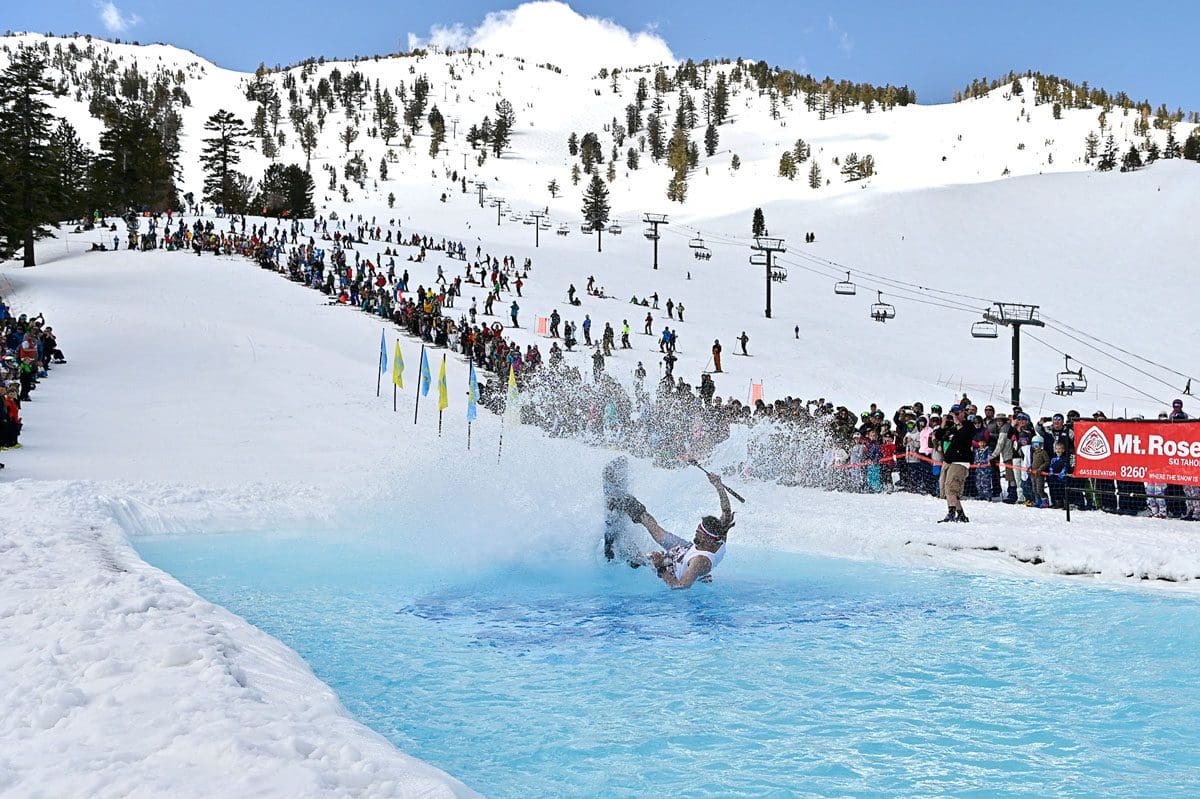Mt. Rose A skier in mid-fall after attempting a pond skim at a snowy mountain resort. The scene is lively with a cheering crowd in the background and ski lifts overhead. A sign on the right reads "Mt. Rose". Tall pines and snowy mountain slopes are visible.