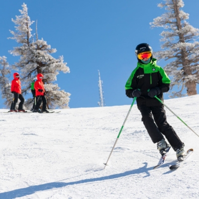 Mt. Rose A skier in a green jacket and black pants glides down a snowy slope under a clear blue sky. Two other skiers in red jackets are visible in the background, standing near snow-covered trees, perhaps taking a break from the engaging kids programs offered at the resort.