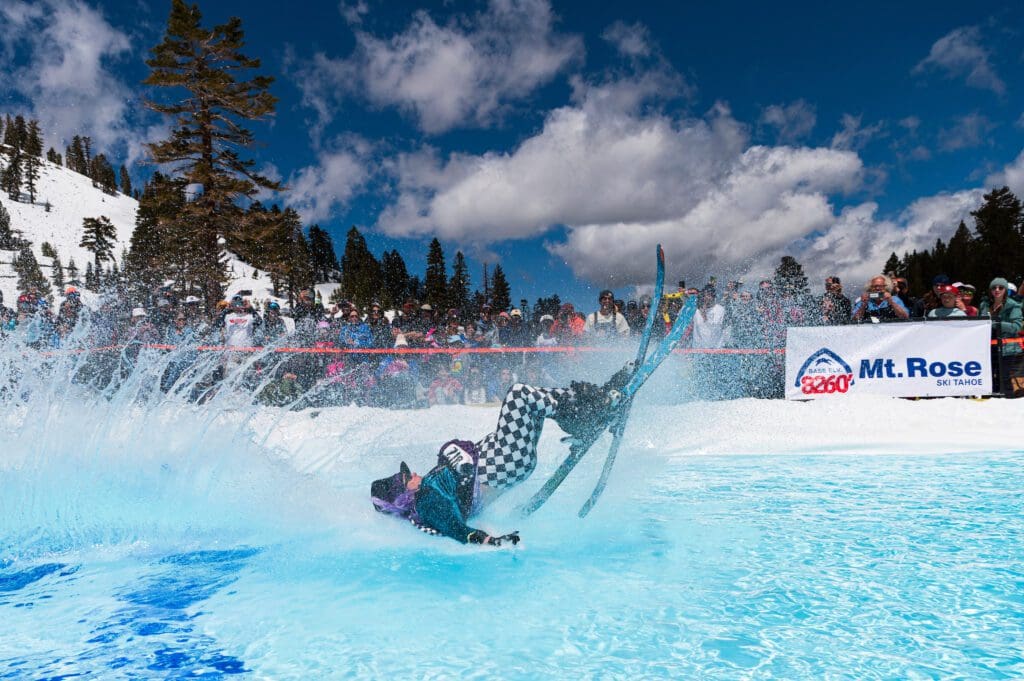Mt. Rose A skier in a checkered outfit crashes into a pool of water during a skiing event, causing a large splash. A crowd watches from the snowy sidelines under a partly cloudy sky. A "Mt. Rose Ski Tahoe" banner is displayed on a nearby fence.