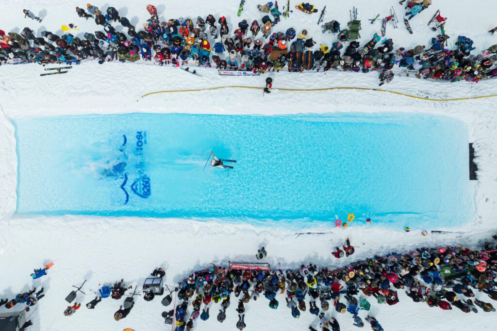 Mt. Rose A large crowd gathers in snowy surroundings to watch a skier in mid air attempting to cross a pool of water. Skis and equipment are scattered around, and people are capturing the event on their cameras. The pool is bright blue, standing out against the white snow.
