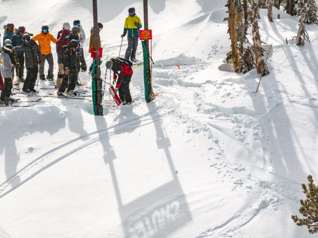 Mt. Rose A group of skiers in colorful winter gear wait in line at a ski lift in a snowy mountain landscape. One skier bends down adjusting equipment near the lift entrance, while others chat and stand on their skis, eager to tackle the challenging chutes ahead. The sun casts clear shadows on the pristine snow.