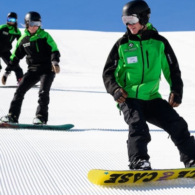 Three snowboarders in green and black outfits glide down a snowy, groomed slope at Ski Tahoe under a clear blue sky. The snowboarder in the front leads the group, while two others follow closely behind, all wearing helmets and goggles. Mt. Rose Ski Tahoe