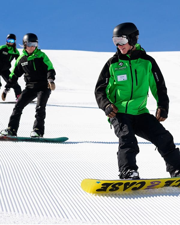 Three snowboarders in green and black outfits glide down a snowy, groomed slope at Ski Tahoe under a clear blue sky. The snowboarder in the front leads the group, while two others follow closely behind, all wearing helmets and goggles. Mt. Rose Ski Tahoe