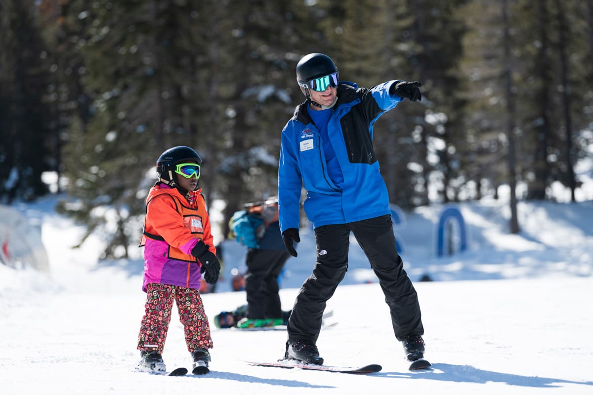 An adult in a blue ski jacket and black helmet points while talking to a child in colorful winter gear and ski equipment on a snowy slope. Both are wearing goggles. Other skiers and trees are visible in the background. Mt. Rose Ski Tahoe