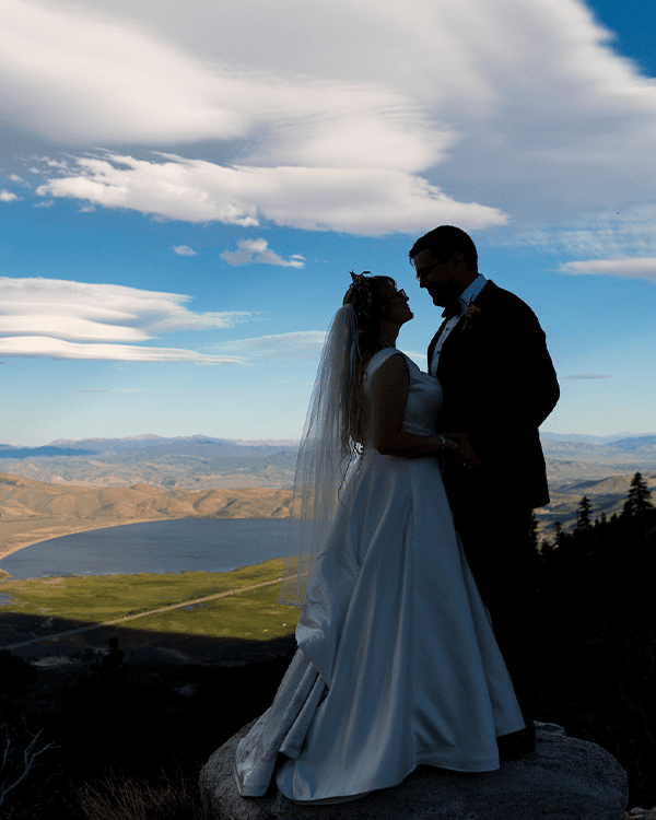 A bride and groom stand closely together on a mountaintop, silhouetted against a picturesque landscape with a vast lake, rolling hills, and a beautiful sky with scattered clouds. The bride wears a white gown and veil, the groom is in a dark suit, and the scene reminiscent of the serene beauty one finds at Ski Tahoe. Mt. Rose Ski Tahoe