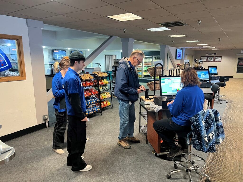 A man stands at a checkout counter staffed by three employees in blue uniforms at a retail store. As they assist him with his purchase, shelves stocked with various snacks can be seen behind them. The store's interior features a carpeted floor and overhead lighting, reminiscent of perusing a dining menu.
 Mt. Rose Ski Tahoe