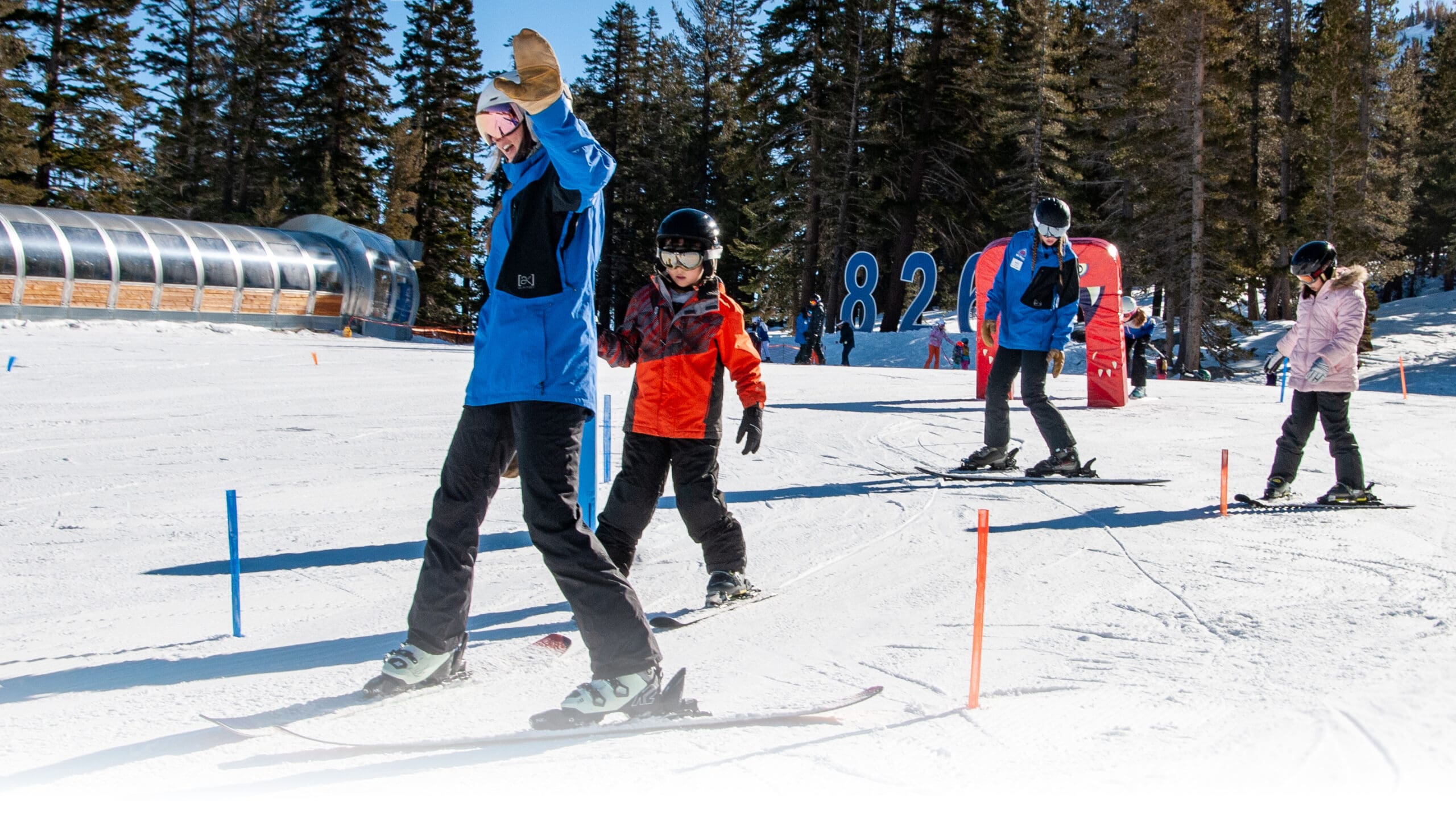 A group of four people, including children and adults, are skiing down a snowy slope on a sunny day. They are all wearing winter gear, including helmets, jackets, and ski boots. There are colorful markers on the snow from the best kids ski lessons, tall pine trees, and a clear blue sky in the background. Mt. Rose Ski Tahoe