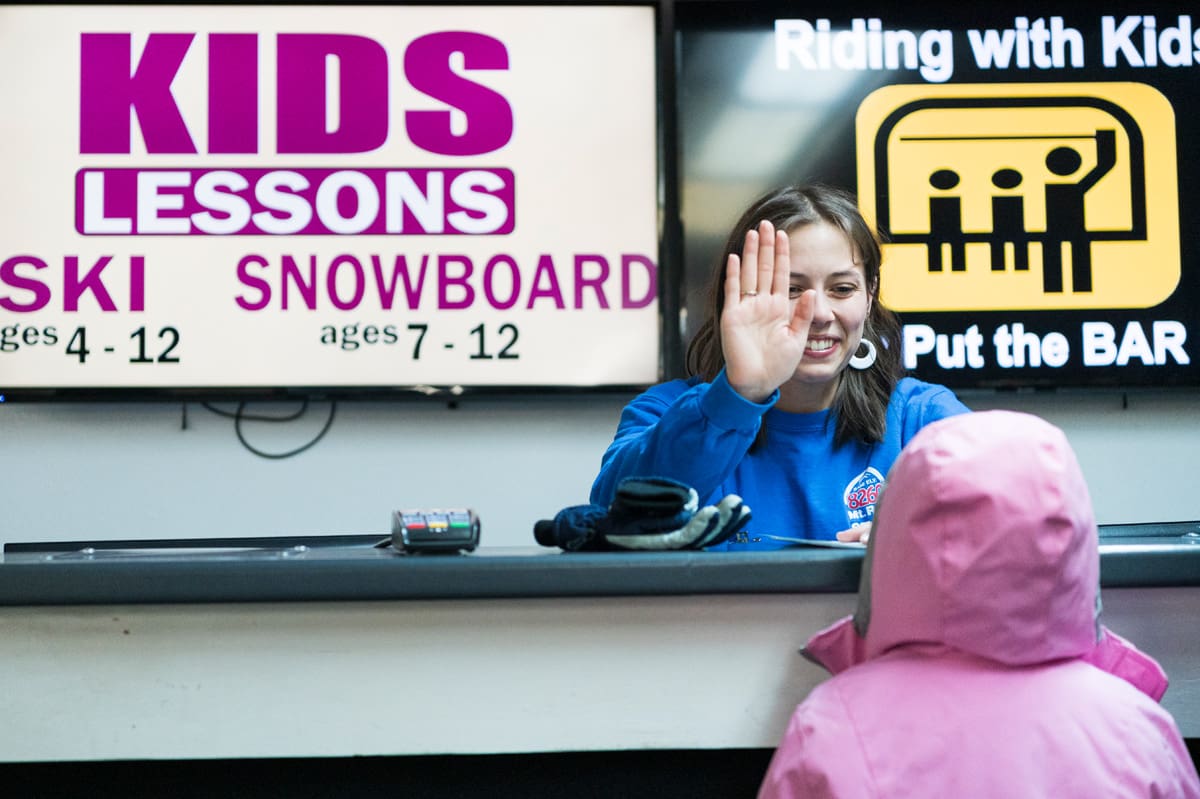 A woman behind a counter is giving a high-five to a child in a pink coat with the hood up. Behind her, there are signs that read "KIDS LESSONS SKI SNOWBOARD" and "Riding with Kids Put the BAR." The environment suggests a ski or snowboarding school setting. Mt. Rose Ski Tahoe