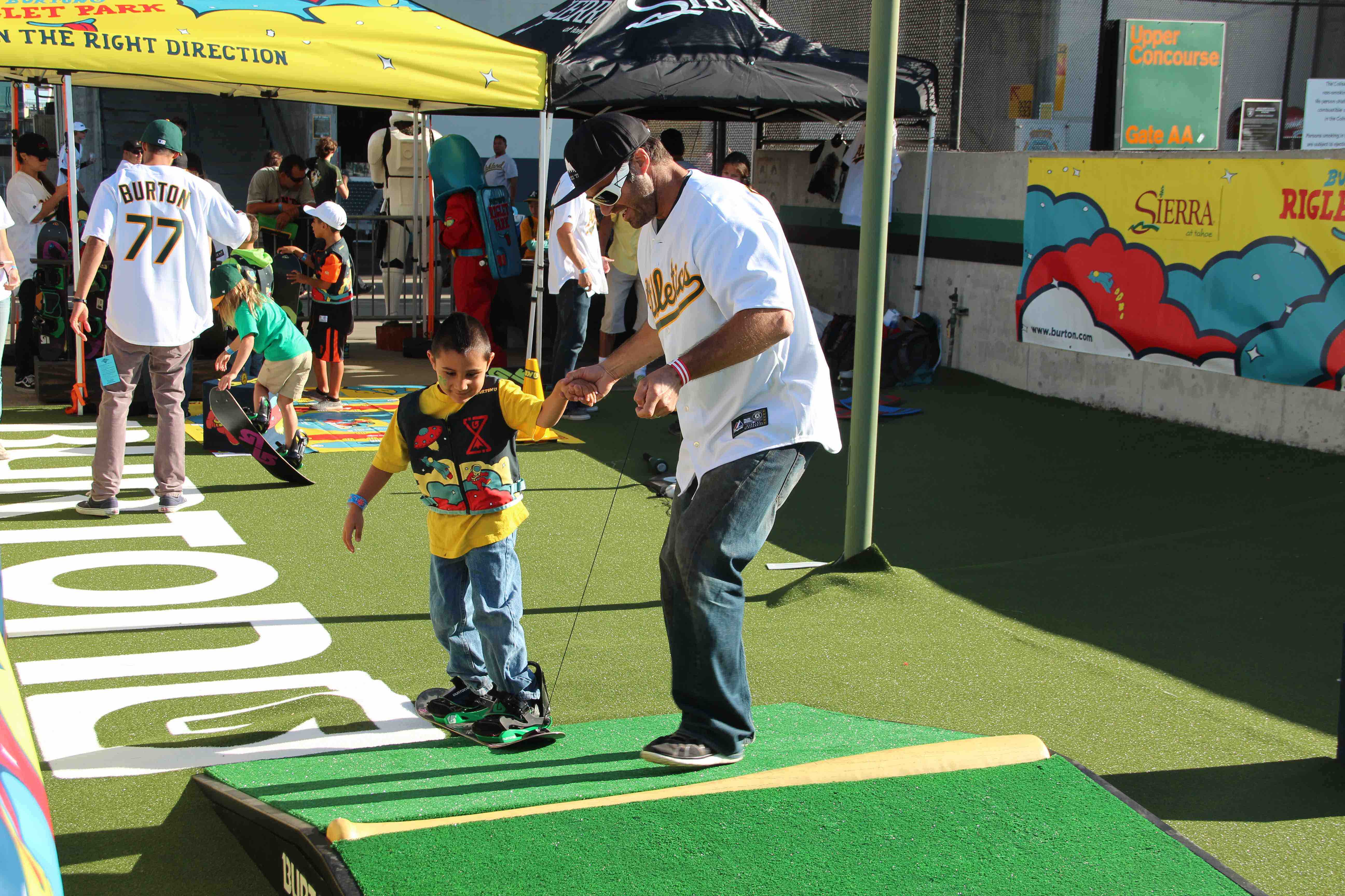 A man helps a young boy balance on a small snowboard on a grassy slope at a promotional event. The area is surrounded by sports-themed decorations and branded tents. Mt. Rose Ski Tahoe