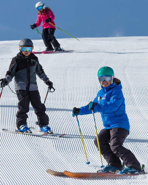 Three people skiing on a sunny, groomed slope at Ski Tahoe. The foreground shows two skiers, one in a blue jacket and green helmet, another in black and gray gear. A third skier in pink descends in the background. All wear helmets and goggles. Mt. Rose Ski Tahoe