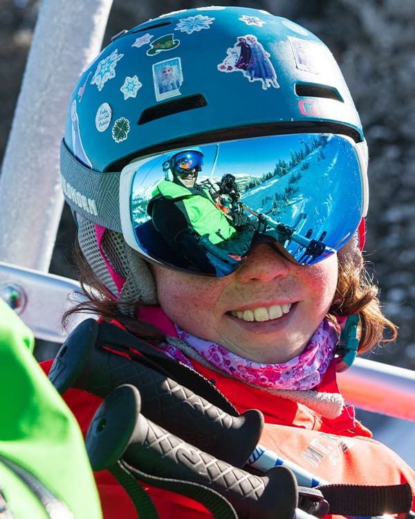 A child wearing a colorful helmet adorned with stickers and reflective ski goggles smiles while sitting on a ski lift at Ski Tahoe. The reflection shows a snowy mountain landscape and another person taking a photo. The child is snug in a red jacket and pink scarf. Mt. Rose Ski Tahoe