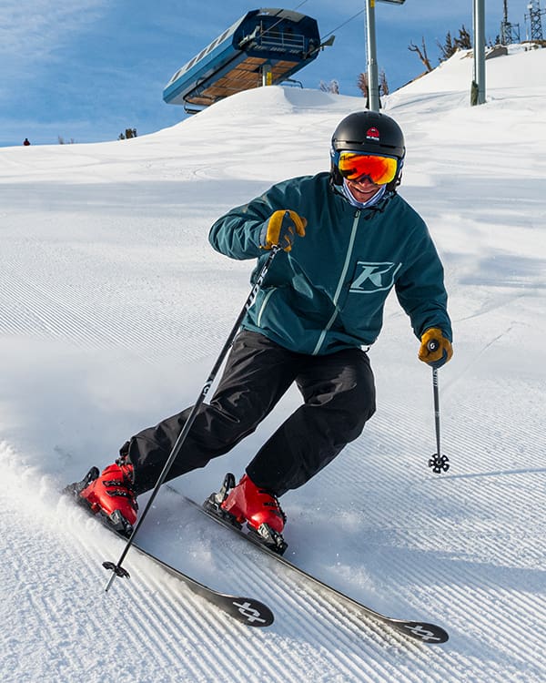 A skier in a green jacket and black pants carves down a groomed Ski Tahoe slope on a sunny day. They wear a helmet, reflective goggles, and red ski boots. A blue ski lift is partially visible in the background against a clear sky. Mt. Rose Ski Tahoe