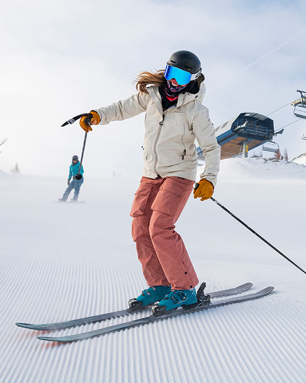 A skier in a white jacket and pink pants gracefully glides down the snowy slope at Ski Tahoe with ski poles, while another person skis in the background. Nearby, a ski lift structure stretches under the partly cloudy sky, capturing the essence of this mountain paradise. Mt. Rose Ski Tahoe