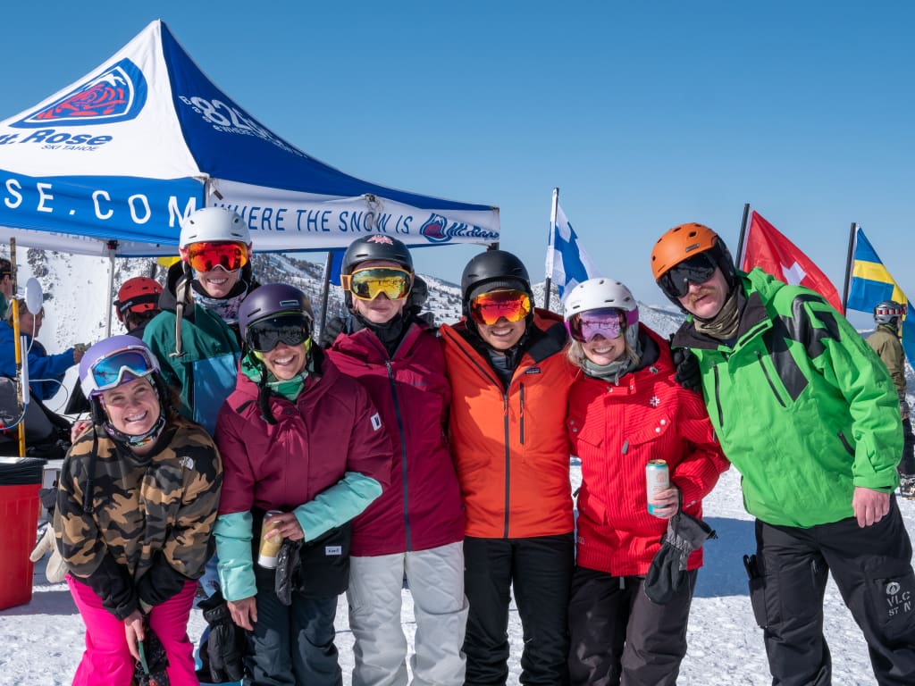 A group of eight people in colorful ski gear stand together smiling in the snow. Behind them is a white tent with a logo and flags, including a Swedish flag. The clear sky suggests a sunny day on the slopes. Mt. Rose Ski Tahoe