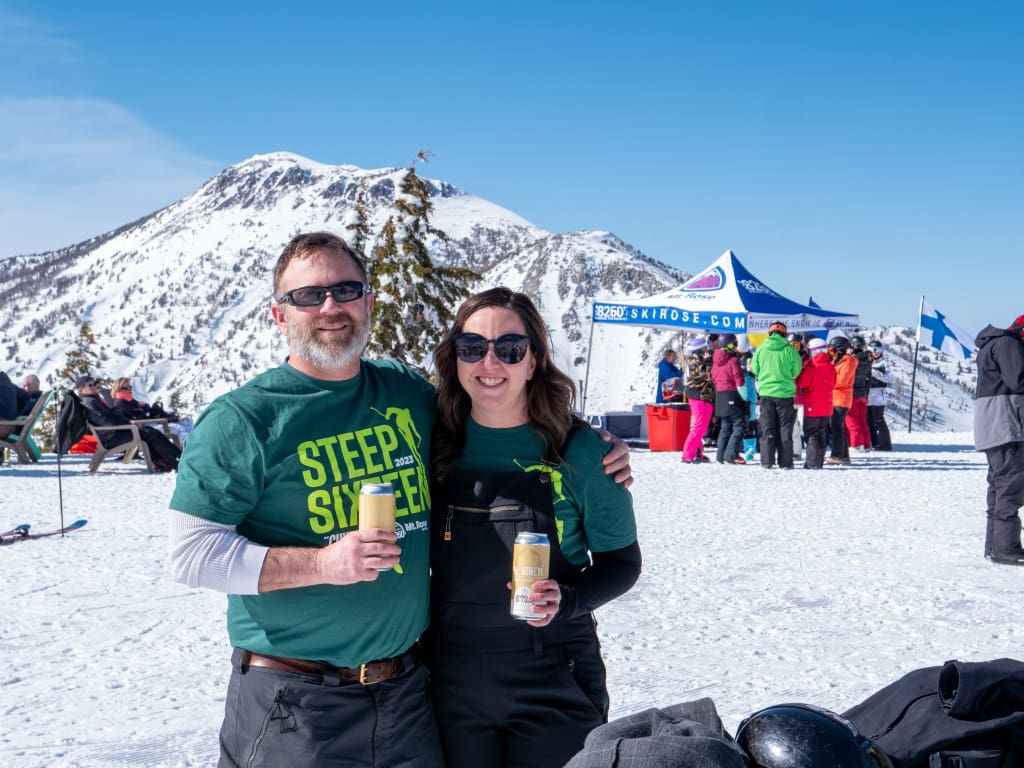A man and a woman in green shirts and sunglasses hold drinks on a snowy mountain. A group of people and a tent are in the background, with a snow-covered peak under a clear blue sky. Mt. Rose Ski Tahoe