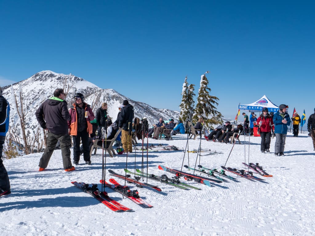 A snowy mountain scene with several people gathered, some standing and others sitting. Skis and poles are lined up on the snow. A tent in the background reads "Demo Center." Clear blue sky and a snow-covered peak complete the view. Mt. Rose Ski Tahoe