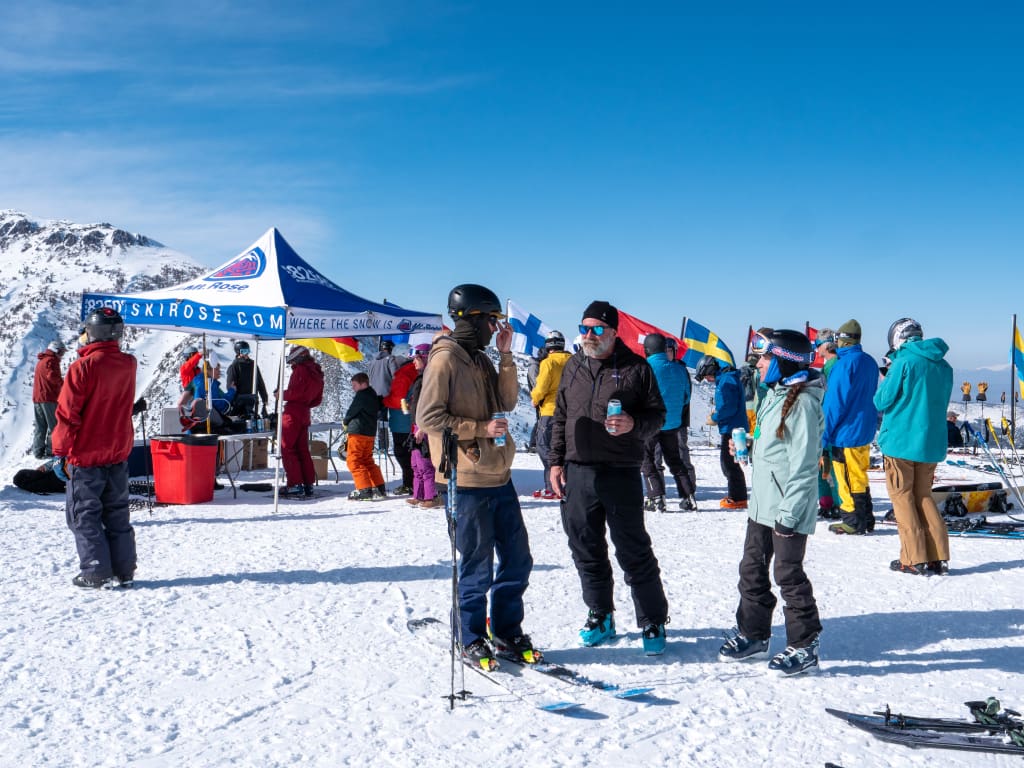 People gather on a snowy hilltop with ski gear, chatting and holding drinks. A branded tent is visible, along with international flags in the background. The sky is clear, highlighting the snow-covered mountains. Mt. Rose Ski Tahoe