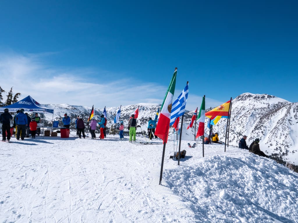 A snowy mountain peak with various national flags, including Italy, Greece, and Colombia, waving in the wind. People in winter gear are standing and interacting near a blue tent. Snow-covered mountains are visible under a clear blue sky. Mt. Rose Ski Tahoe