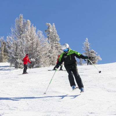 A person in a green and black jacket skis downhill on a snowy slope under a clear blue sky. Another person in a red jacket stands in the distance with snowy trees surrounding them. Mt. Rose Ski Tahoe