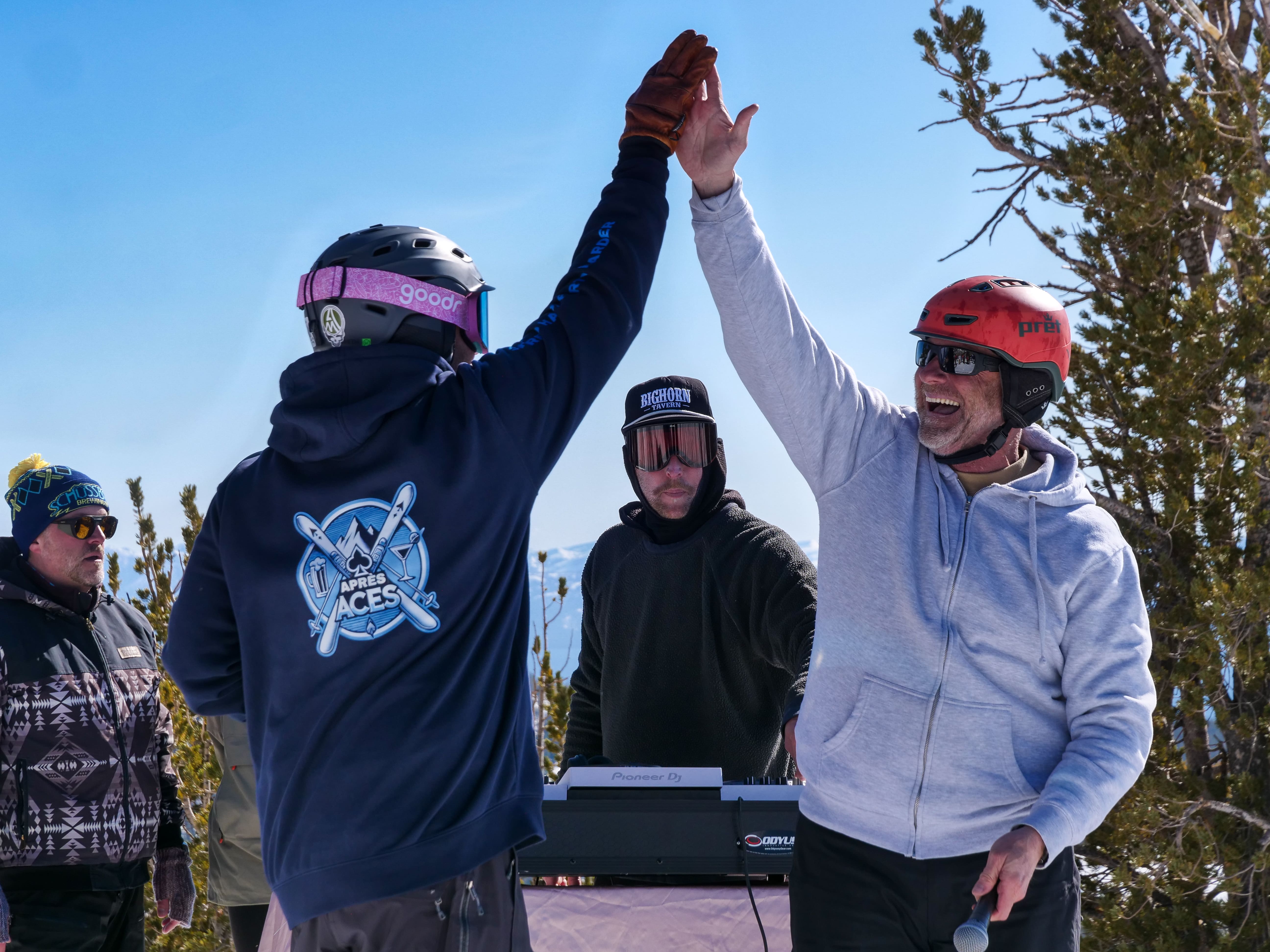 Two people wearing helmets and winter jackets high-five outdoors in a snowy, mountainous area while others stand nearby. One plays a keyboard. The mood is celebratory and energetic. Mt. Rose Ski Tahoe