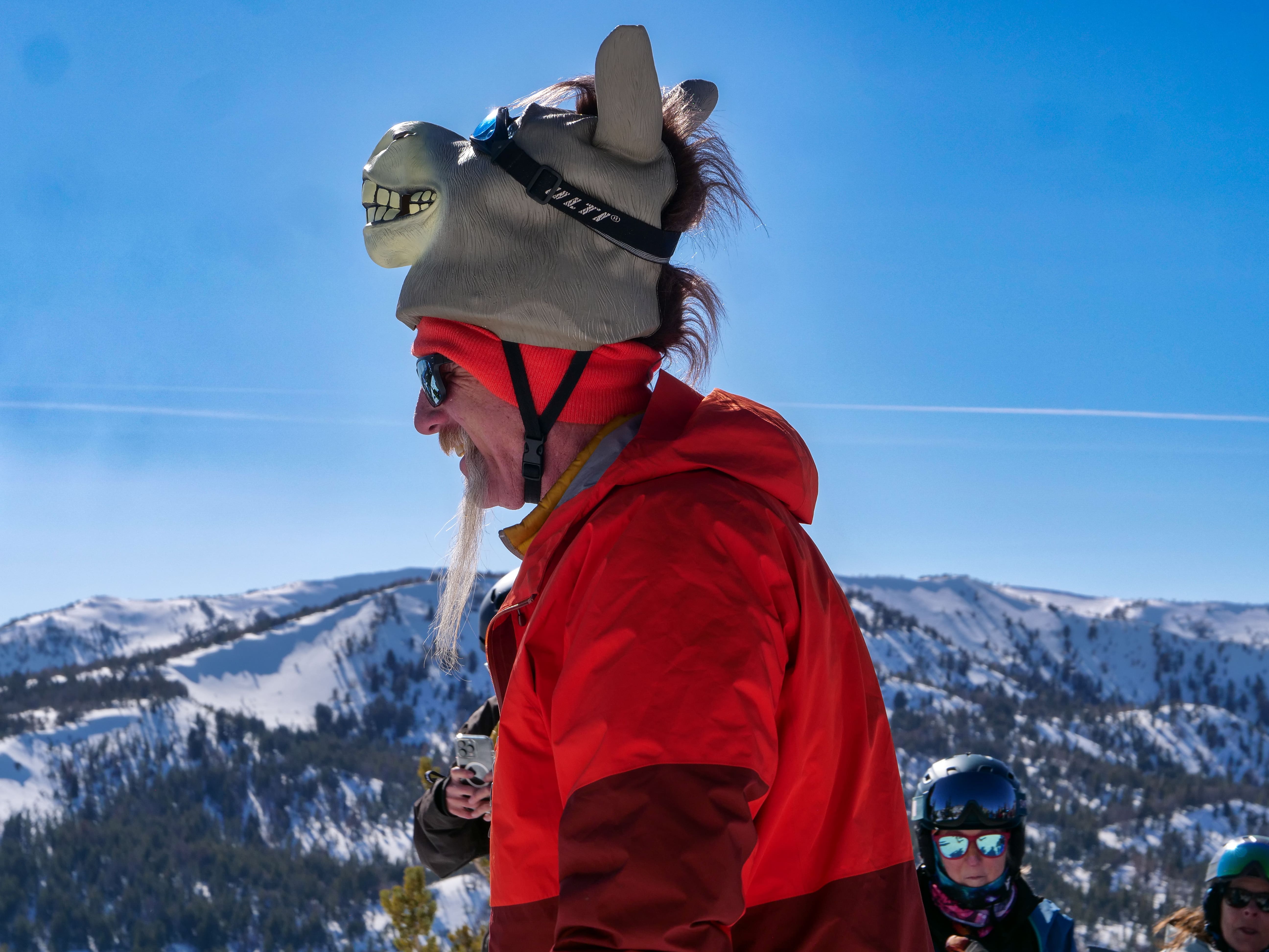 A person in a red jacket and sunglasses wears a whimsical donkey hat with ears and teeth, standing outdoors in a snowy mountain landscape. Other people in winter gear are visible in the background. Mt. Rose Ski Tahoe