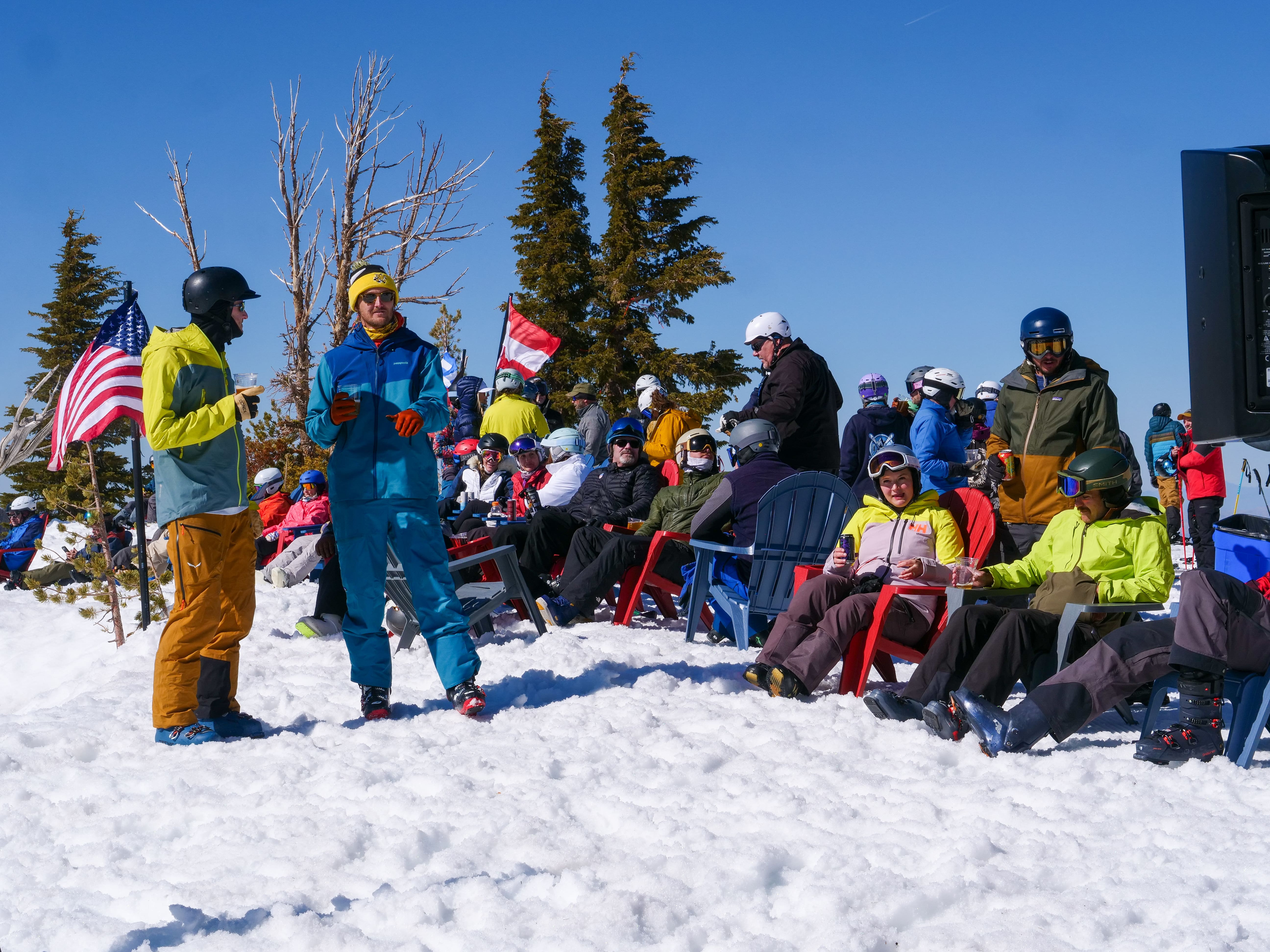 A group of people in ski gear sit and stand on the snowy ground under a blue sky, some holding American flags, while others relax in chairs near evergreen trees. Mt. Rose Ski Tahoe
