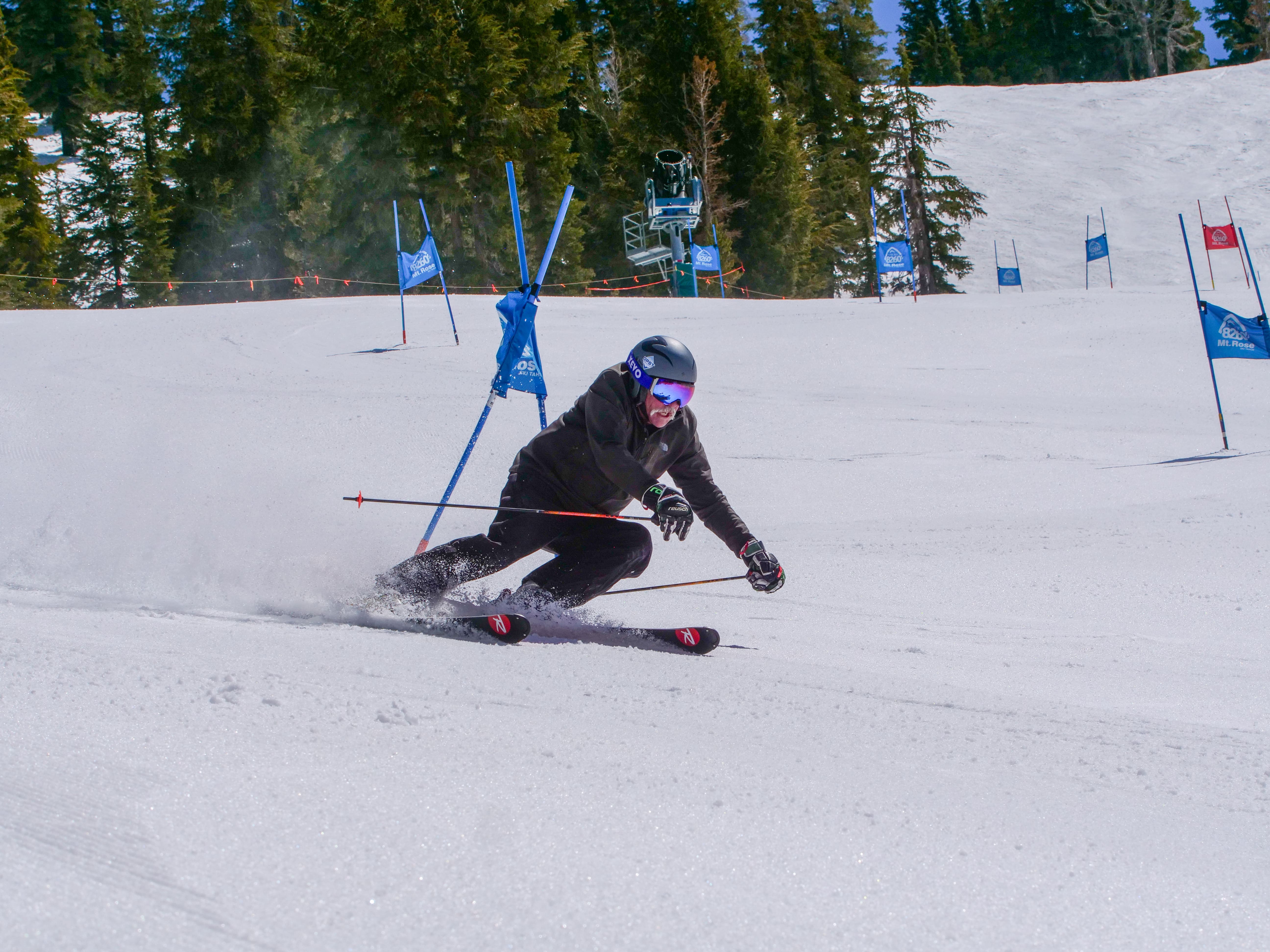 A skier wearing a helmet and goggles makes a sharp turn on a snowy downhill slalom course, passing blue gates with trees and a chairlift visible in the background. Mt. Rose Ski Tahoe