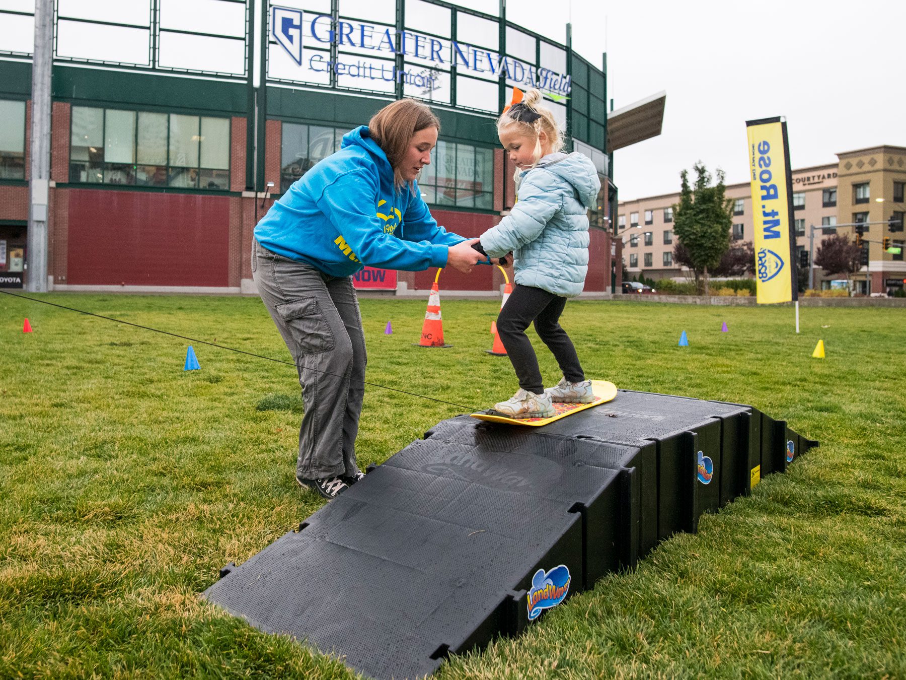 A woman helps a young girl balance on a small ramp while standing on a board, outdoors on grass near buildings and event signs. Mt. Rose Ski Tahoe