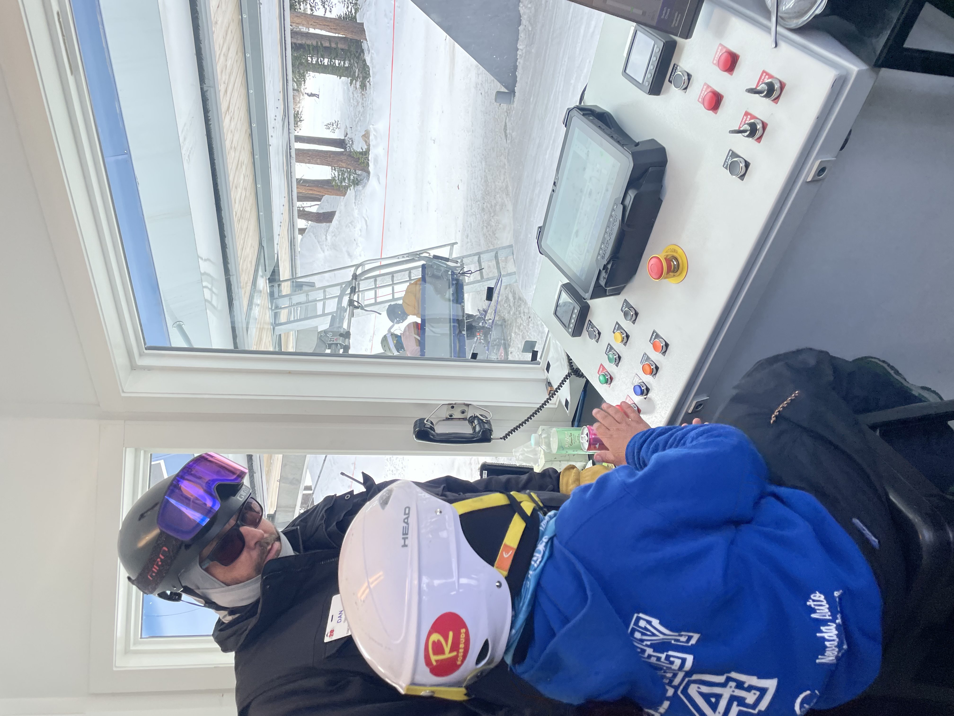 An adult in ski gear supervises a child wearing a helmet and blue hoodie as they sit at a ski lift control panel indoors, with snowy slopes and skiers visible through the window. Mt. Rose Ski Tahoe