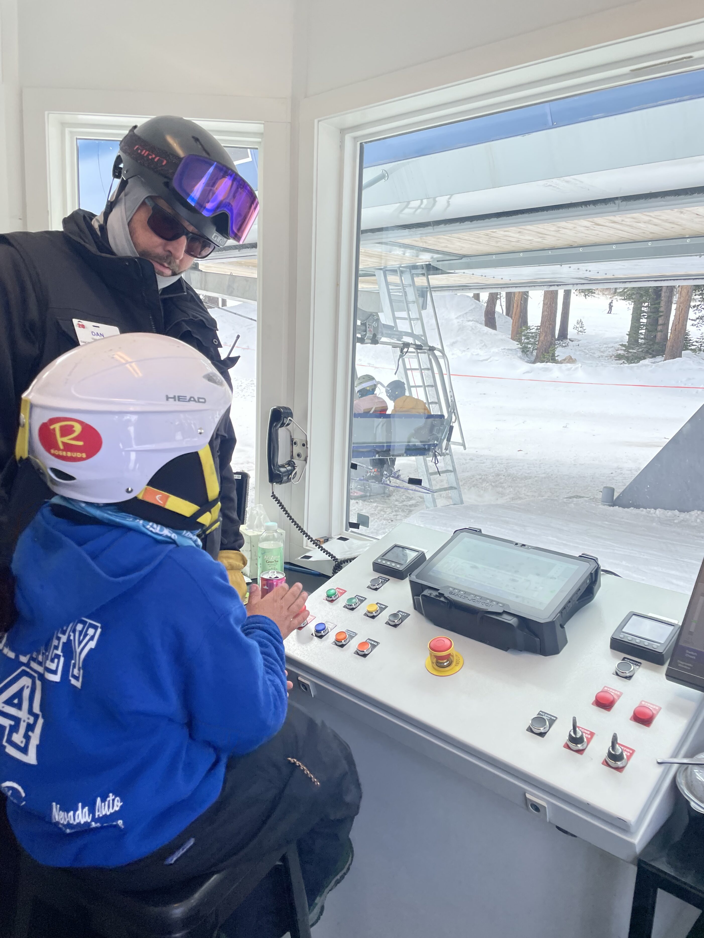 A ski instructor in red stands on snowy ground, talking to four students with helmets and winter gear who are sitting or kneeling on the snow, surrounded by tall pine trees. Mt. Rose Ski Tahoe