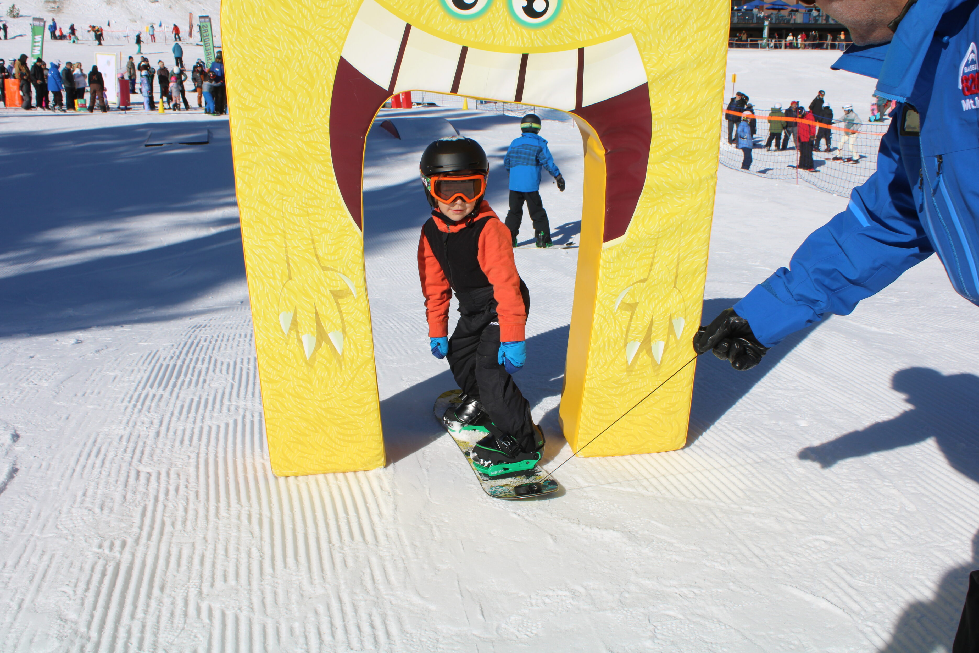A young child wearing a helmet and goggles snowboards through a yellow archway with cartoon teeth and eyes on a snowy ski slope, while an adult in blue holds a guiding rope. People and ski equipment are visible in the background. Mt. Rose Ski Tahoe