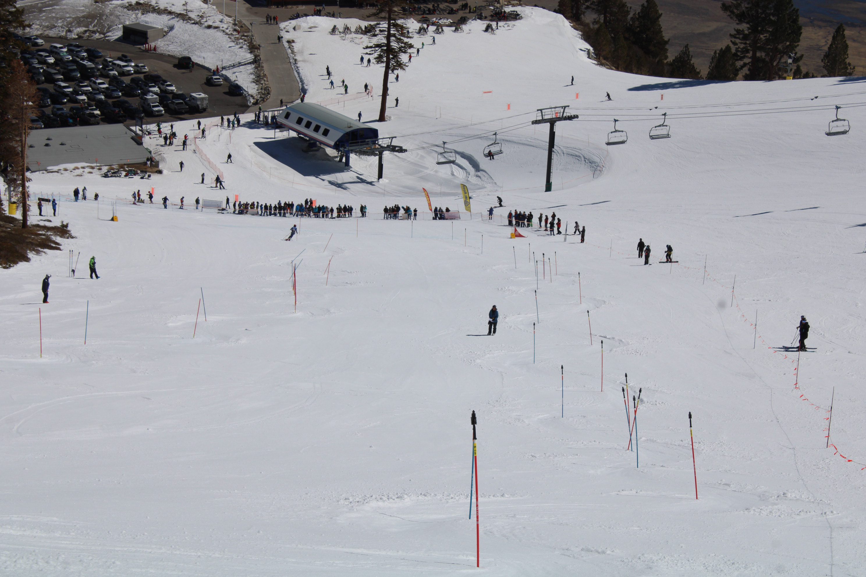 A snowy ski slope with skiers descending, lift chairs overhead, and people waiting in line near a ski lift station. Trees and parked cars are visible in the background. Mt. Rose Ski Tahoe