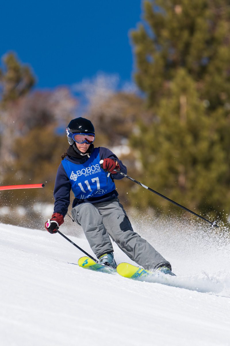 A skier wearing a blue bib and helmet races downhill on snowy slopes, kicking up powder. Trees and a clear blue sky are visible in the background. Mt. Rose Ski Tahoe