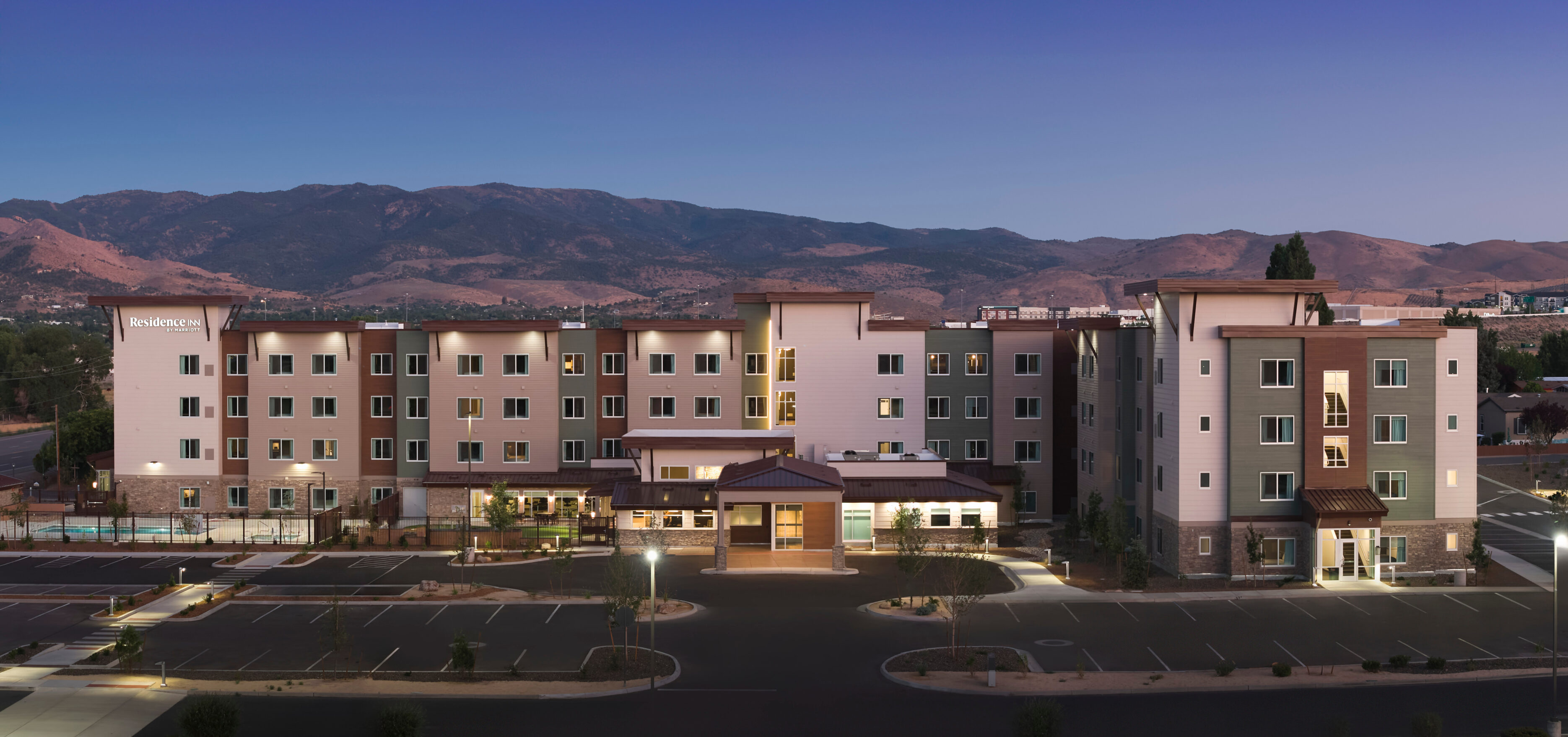 A modern, multi-story hotel building with lit windows sits in front of a mountainous landscape at dusk, offering perfect ski trip lodging for family ski trips, with an empty parking lot in the foreground. Mt. Rose Ski Tahoe