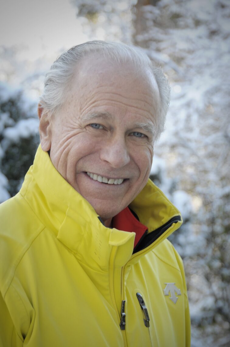 An older man with gray hair smiles outdoors, wearing a bright yellow jacket at a film festival. Snow-covered trees and bushes are visible in the softly focused background, suggesting a winter setting. Mt. Rose Ski Tahoe