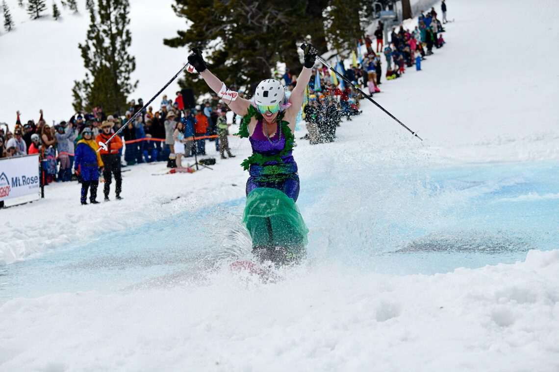 A skier in a mermaid costume splashes through a water obstacle on a snowy slope, arms raised and surrounded by cheering spectators during a winter event. Mt. Rose Ski Tahoe