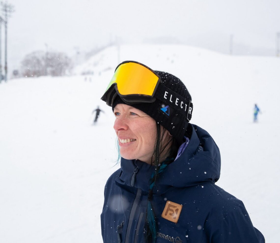 A woman wearing a black ski hat, yellow ski goggles, and a blue jacket smiles while standing on a snowy slope, as if ready for a film festival set in the mountains, with skiers in the background on a cloudy, snowy day. Mt. Rose Ski Tahoe