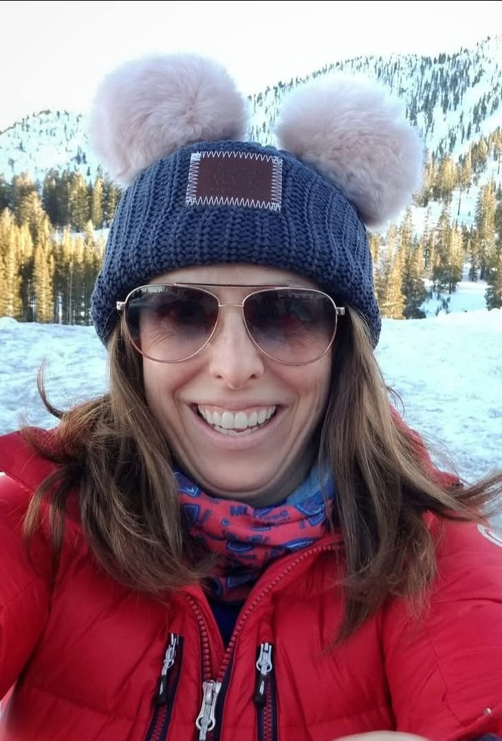 A woman wearing sunglasses, a red winter jacket, and a blue knit hat with two fluffy pom-poms smiles outdoors in a snowy mountain landscape, capturing the spirit of a film festival set against breathtaking alpine scenery. Mt. Rose Ski Tahoe