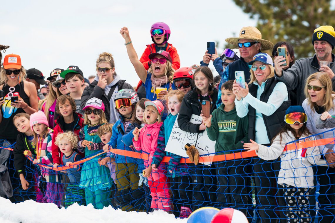 A group of excited children and adults in winter clothing stand behind a blue netting, cheering and taking photos at an outdoor snowy event under a partly cloudy sky. Mt. Rose Ski Tahoe