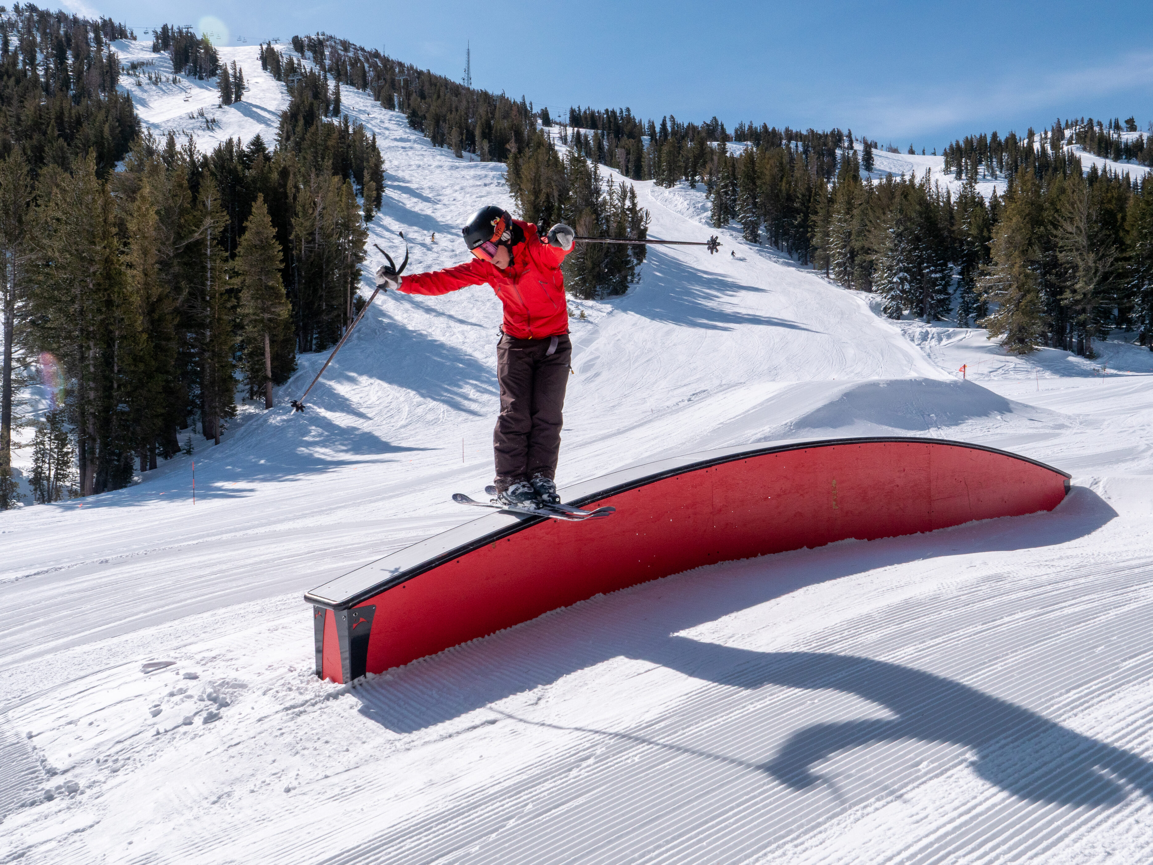 A snowboarder in a red jacket and black helmet balances on a curved red rail in a snowy terrain park, surrounded by pine trees and mountains under a clear blue sky. Mt. Rose Ski Tahoe