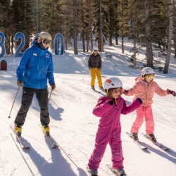 Two adults and two children wearing helmets and ski gear ski down a snowy Ski Tahoe slope among tall trees. The children, in bright pink suits, lead the way. Large blue numbers, 8200, are visible in the background. Mt. Rose Ski Tahoe