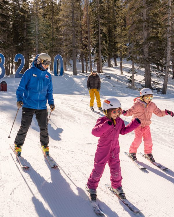 Two adults and two children wearing helmets and ski gear ski down a snowy Ski Tahoe slope among tall trees. The children, in bright pink suits, lead the way. Large blue numbers, 8200, are visible in the background. Mt. Rose Ski Tahoe